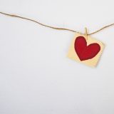 Marketing mit Herz – Eine braune Schnur hängt an einer weißen Wand. Mit einer kleinen Klammer wurde ein Zettel mit einem gemalten rotem Herz an die Schnur festgesteckt.