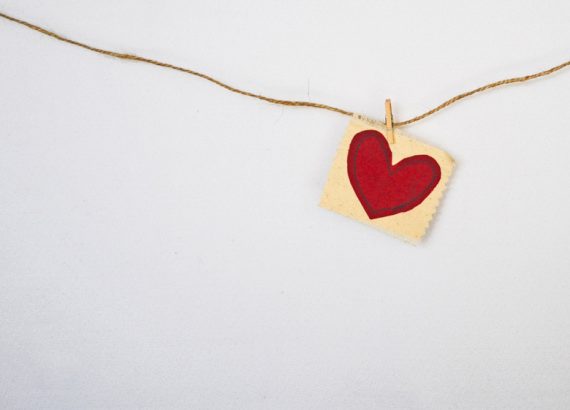 Marketing mit Herz – Eine braune Schnur hängt an einer weißen Wand. Mit einer kleinen Klammer wurde ein Zettel mit einem gemalten rotem Herz an die Schnur festgesteckt.
