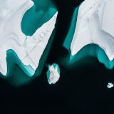 Drei Eisberge sind frontal von oben fotografiert. Das Wasser wirkt fast schwarz und färbt sich nur an den Stellen Türkis, wo der Eisberg im Wasser liegt.