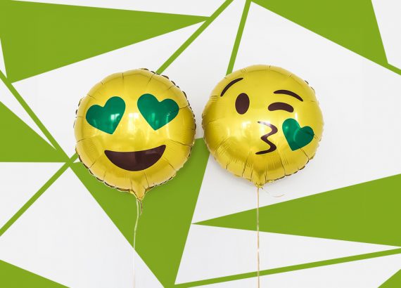 Es sind zwei Luftballon mit Emoji-Motiven. Der linke Luftballon ist rund, gelb und das Emoji hat einen offenes lachen. Die Augen sind zwei grüne Herzen. Der rechte Luftballon hat ein Kussmund geformt woraus ein grünes Herz kommt. Die Augen zwinkern einem zu. Diese Smiley werden im Emoji-Marketing eingesetzt.