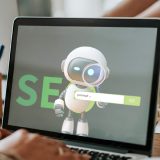 Ein aufgeklappter Laptop, auf dem Bildschirm steht das Wort SEO und es ist ein kleiner Roboter mit einer Lupe in der Hand zu sehen